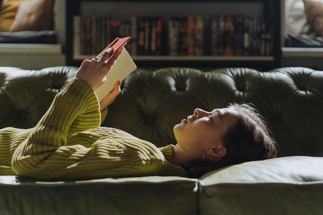 gallery-5 A young woman lies on a sofa reading a book, enjoying a peaceful indoor moment.