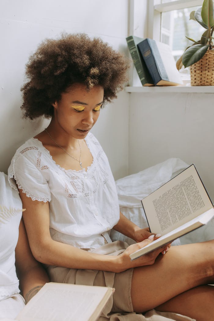 gallery-6 A young woman with natural hair enjoys reading a book by the window with warm sunlight.
