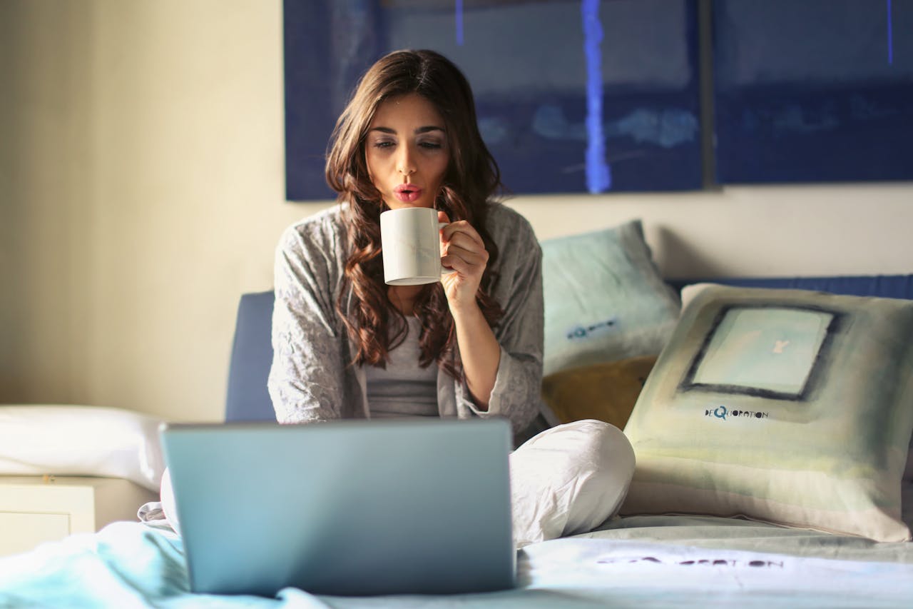 gallery-1 A woman enjoying coffee while working from home in a cozy bedroom setting with a laptop.